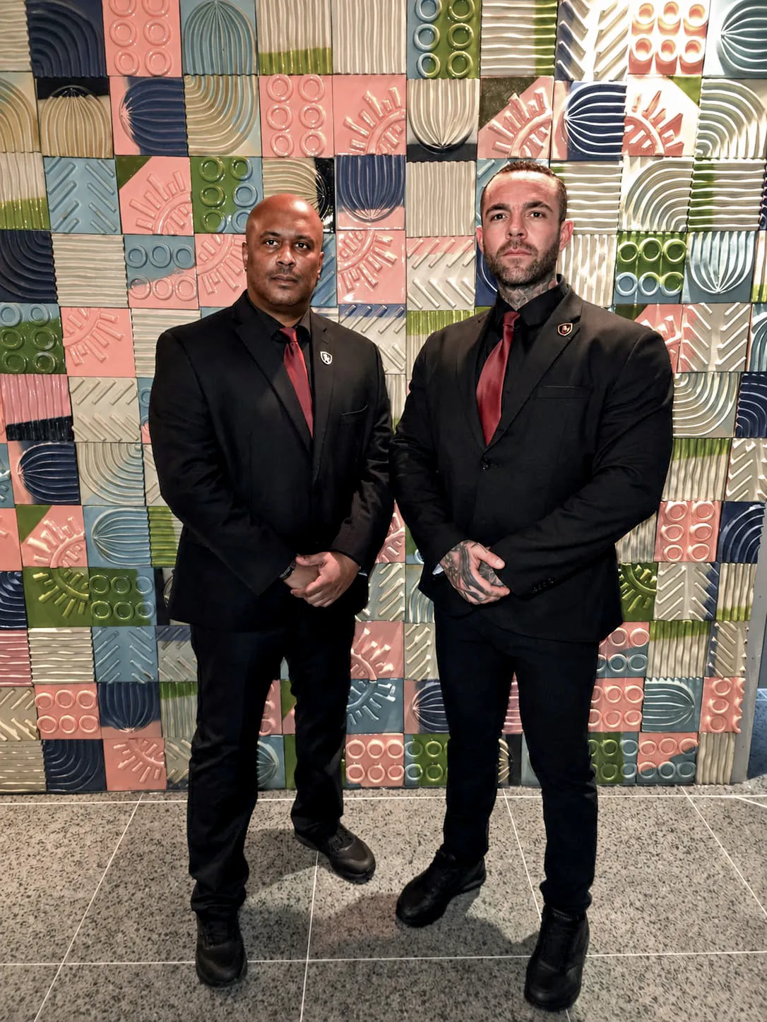 Two Prestige Guard officers in black suits and burgundy ties on post in front of a decorative tiled wall